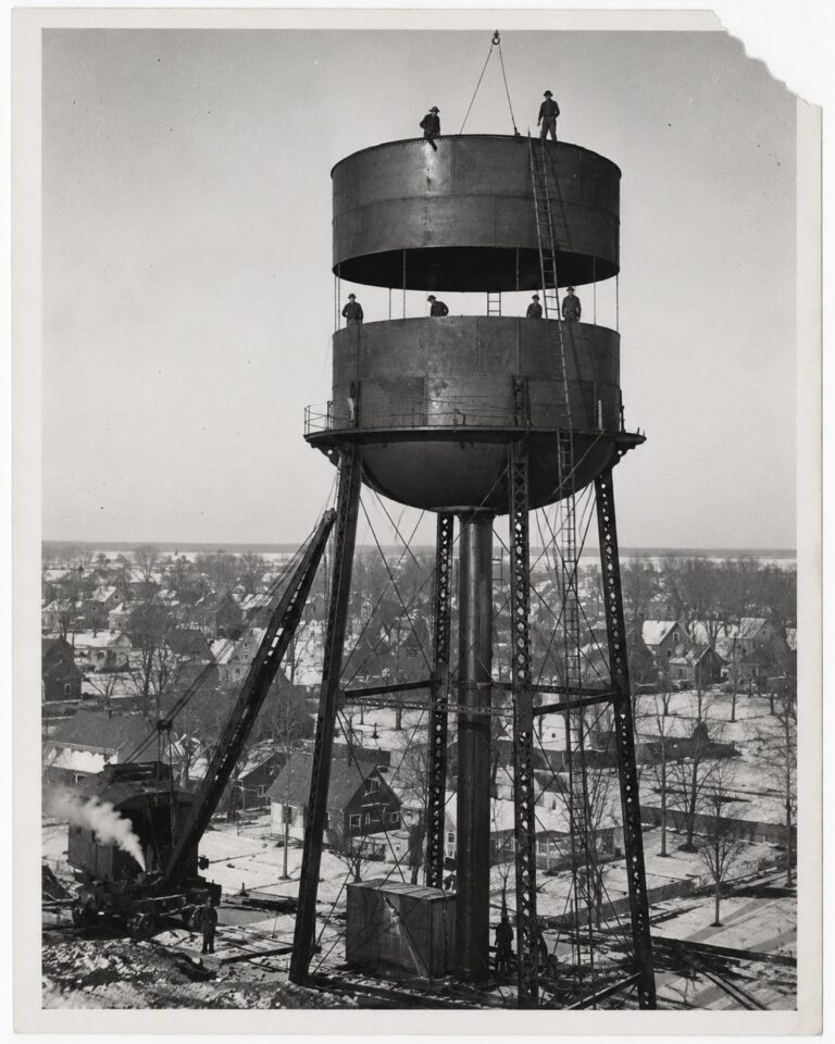 Workers on the half-assembled steel water tower above the rooftops, circa 1930