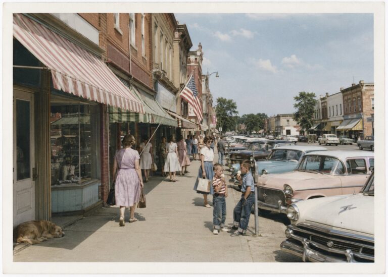 Main Street with striped awnings, parked cars and shoppers on a summer Saturday, circa 1958