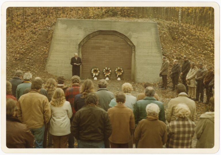 Townspeople with heads bowed before the sealed tunnel portal, three wreaths and three hard hats at its base, September 1974